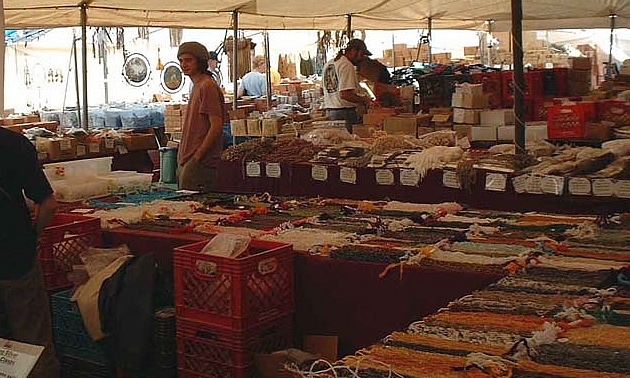 A man stands in an outdoor market, with tables full of handmade crafts.