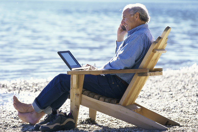 An elderly man sitting in a wooden beach chair talking on his cell phone.