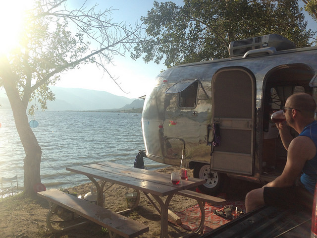 Dave Barton sitting in a lawn chair outside of his vintage airstream.  The airstream is parked on the edge of a lake