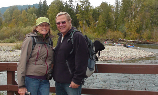 Liz and Anders stand in front of a rail. Behind them is the curve of a river, and several kayaks.