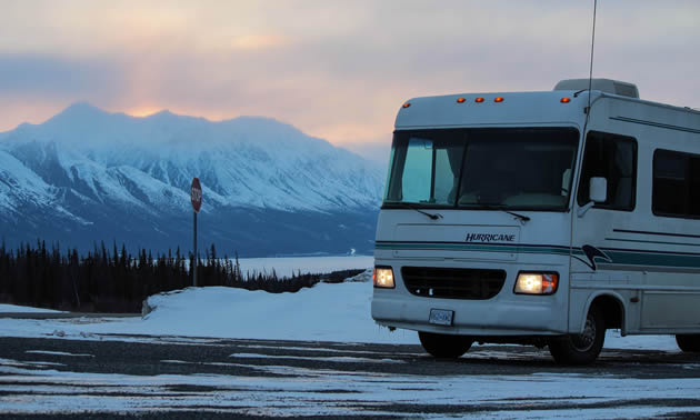 An RV on snowy road with snowy mountains in the background. 