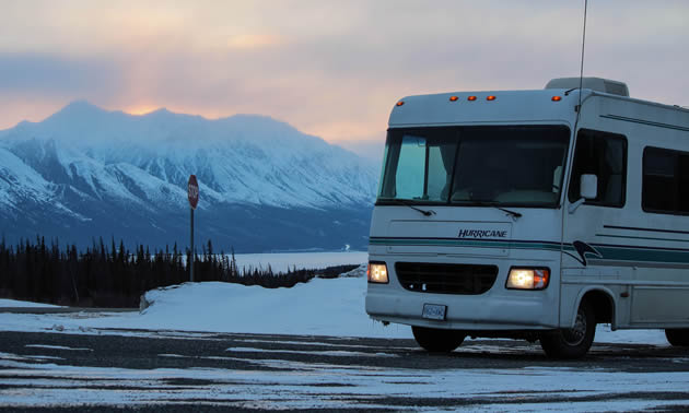 An RV on snowy road with snowy mountains in the background.