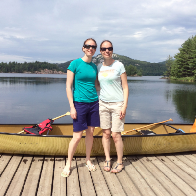 Two women wearing sunglasses, T-shirts, shorts and sandals smile in front of a backdrop that includes a yellow canoe, a lake, distant mountains and cloudy skies.