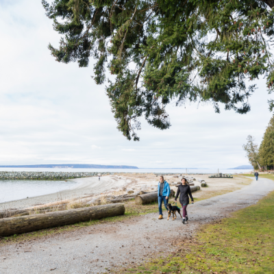 Two women take a dog for a walk along a shoreline.