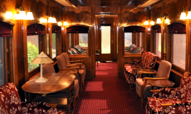 An old train car has wooden walls and a maroon carpet. It is furnished with beige and dark red chairs.