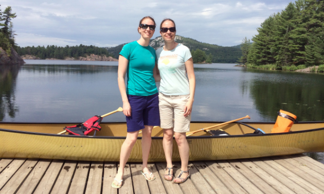 Two women wearing sunglasses, T-shirts, shorts and sandals smile in front of a backdrop that includes a yellow canoe, a lake, distant mountains and cloudy skies.