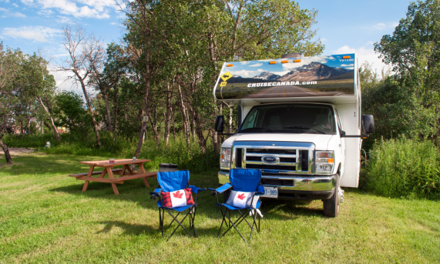 Two blue fold-out chairs face towards the camera with pillows that emblazon the Canada flag. A picnic table and RV are behind the chairs in a pleasant-looking green park area.