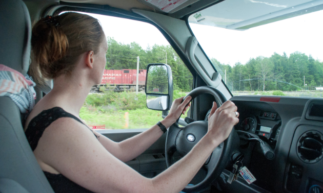 A woman drives an RV and takes her eyes off the road for a moment to look upon a Canadian fixture: a red Canadian Pacific train travelling on a parallel course.