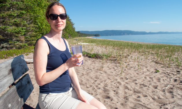 A woman wearing sunglasses, a tank top and shorts holds a beverage on a beach.