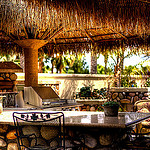 A picture of an outdoor area to eat. There is a thatched roof, a table with 2 iron chairs, and a BBQ in the background.