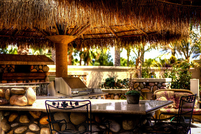 A picture of an outdoor area to eat. There is a thatched roof, a table with 2 iron chairs, and a BBQ in the background.