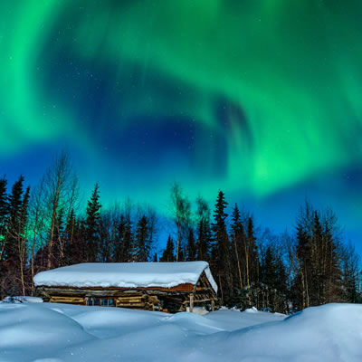 A cabin is buried in deep snow with northern lights in the sky. 