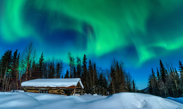 A cabin is buried in deep snow with northern lights in the sky. 