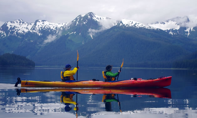 Two people are kayaking, with snowy mountains in the background. 