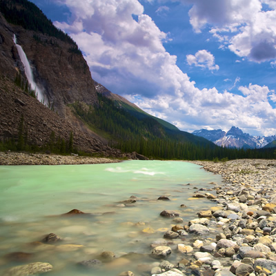 Picture of green-hued stream, with waterfall and mountains in background. 