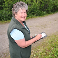 Rose, wearing a vest, jeans and t-shirt, smiles at the camera while she takes notes on a clipboard.