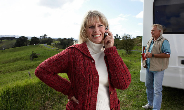 A smiling women pictured in the forground is talking on her cell phone while her husband, who is seen in the backround, is holding a cup of coffee next to their RV.