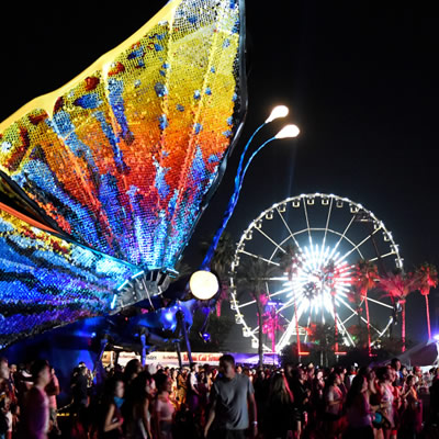 Large group of festival goers at night, with lit-up Ferris wheel in background and large, colourful illuminated butterfly sculpture in foreground. 