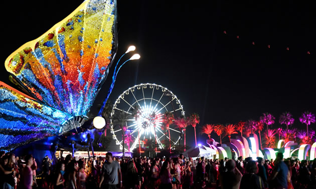 Large group of festival goers at night, with lit-up Ferris wheel in background and large, colourful illuminated butterfly sculpture in foreground. 