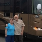 A man stands with his arm around his wife who is wearing a blue shirt in from of a large RV in shades of brown.