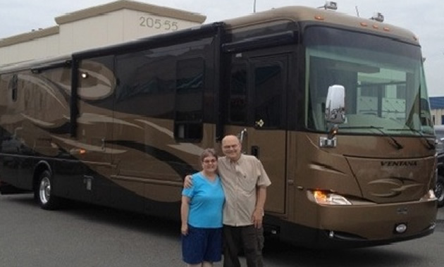 A man stands with his arm around his wife who is wearing a blue shirt in from of a large RV in shades of brown.