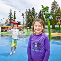 A little boy and girl smile at the camera from  brightly coloured spray park.