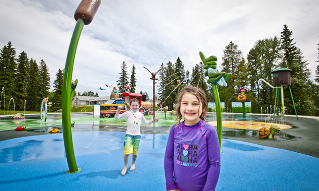 A little boy and girl smile at the camera from  brightly coloured spray park.