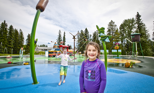 A little boy and girl smile at the camera from brightly coloured spray park.
