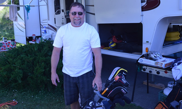 Smiling man in sunglasses, with golf clubs at hand and an RV in the background