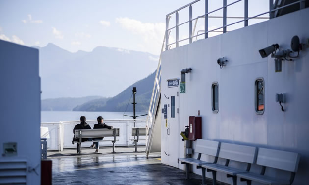 Two people sitting on bench on deck of ferry. 