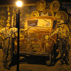 a painted mural on the side of a building at night lit by a vintage streetlight; mural is of two men standing beside an old truck