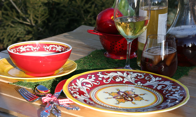 Attractive red and yellow designs on china is set out in front of ice tea, an apple, and glasses of wine to create an appealing camp setting.