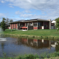 An attractive dark red building stands beneath a blue sky, on green grass and above a pond and fountain.