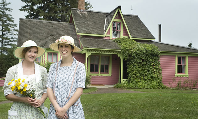 Hisotric house, with two ladies dressed in period costume in the foreground, one of them holding a bouquet of yellow daisies.