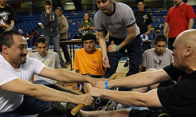 Two men sitting with legs outstretched, and holding onto stick with both hands. 