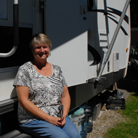 A woman sits on the steps leading into her white and black RV. A water dispenser is beside the entrance.