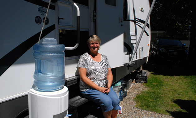 A woman sits on the steps leading into her white and black RV. A water dispenser is beside the entrance.