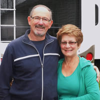 A couple stand, arms around each other, smiling for the camera in front of their RV.