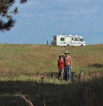 A photo of Kevin and Ruth Read in the forground, and Sherman their motorhome on a grassy hill in the background.