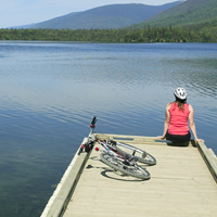 A woman sits on a dock looking over a lake with a bike beside her.