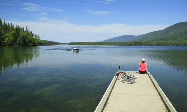 A woman sits on a dock looking over a lake with a bike beside her.