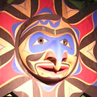 A large decorative carved native mask resembles a sun with a central face and arms that look like birds. It is painted in reds, purples and browns.