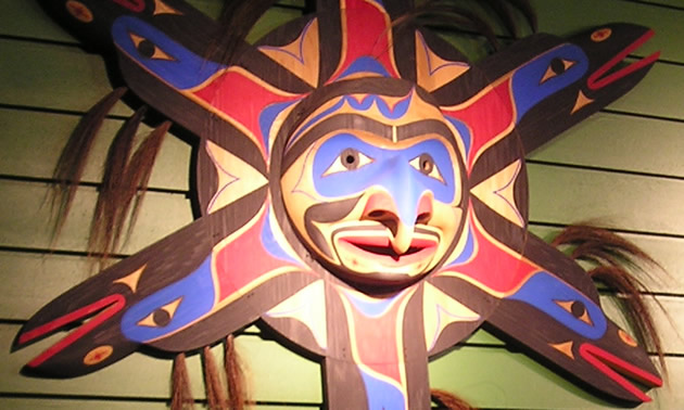 A large decorative carved native mask resembles a sun with a central face and arms that look like birds. It is painted in reds, purples and browns.