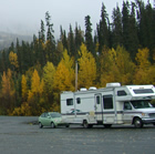 Rae's RV, hauling a green hatchback, is parked to the right against yellow autumn trees and a misty mountain background.