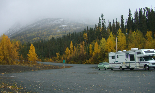 Rae's RV, hauling a green hatchback, is parked to the right against yellow autumn trees and a misty mountain background.