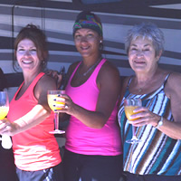 A row of smiling women wearing casual summer clothing and holding out stemmed glasses of orange liquid