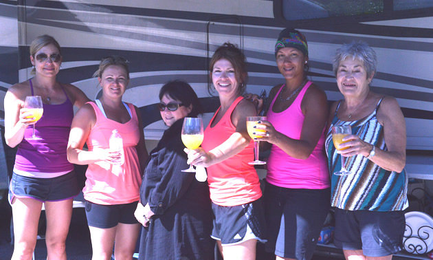 A row of smiling women wearing casual summer clothing and holding out stemmed glasses of orange liquid