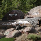 Natural rounded boulders and rocks bear the words Myrtle Creek; a log lying in front of the rocks bears the words 