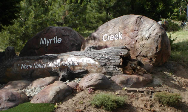 Natural rounded boulders and rocks bear the words Myrtle Creek; a log lying in front of the rocks bears the words 