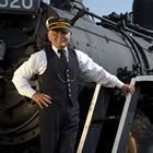 A train conductor stands posed on the front of an antique engine.